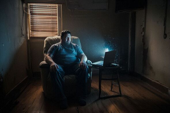 A man sits in a dim apartment lit only by a laptop screen, symbolizing isolation and emotional weight.